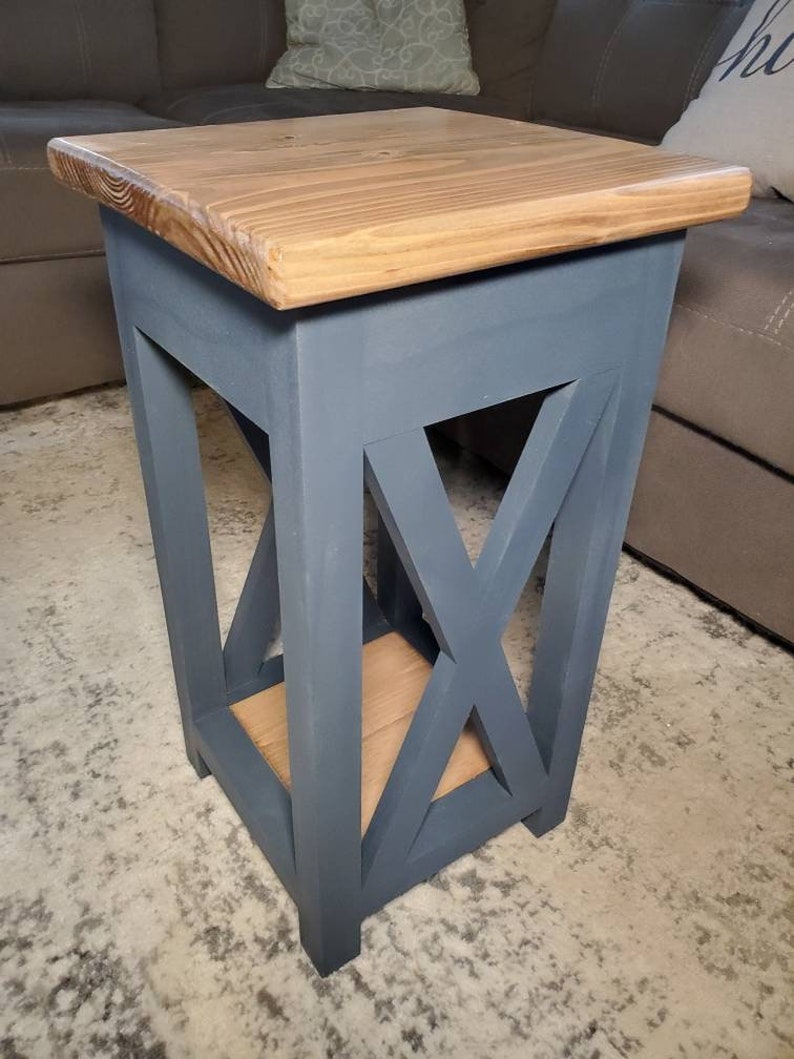 May include: A small, blue, wooden end table with a natural wood top and an X-shaped design on the front. The table is sitting on a gray carpet.