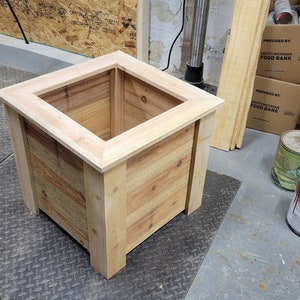 K&ouml;nnte beinhalten: Ein quadratischer Holz-Pflanzkasten mit einer nat&uuml;rlichen Holz-Oberfl&auml;che. Der Pflanzkasten besteht aus unbehandelten Holzbrettern und hat einen erh&ouml;hten Rand oben.