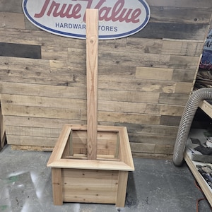 May include: A wooden planter box with a tall, vertical wooden post in the center. The planter box is made of unfinished wood and has a square shape. The post is also made of unfinished wood and is taller than the planter box. The planter box is sitting on a wooden floor.