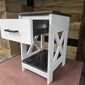 May include: White nightstand with a black top and a single drawer. The drawer is open and has a black pull handle. The nightstand has an X-shaped design on the front.