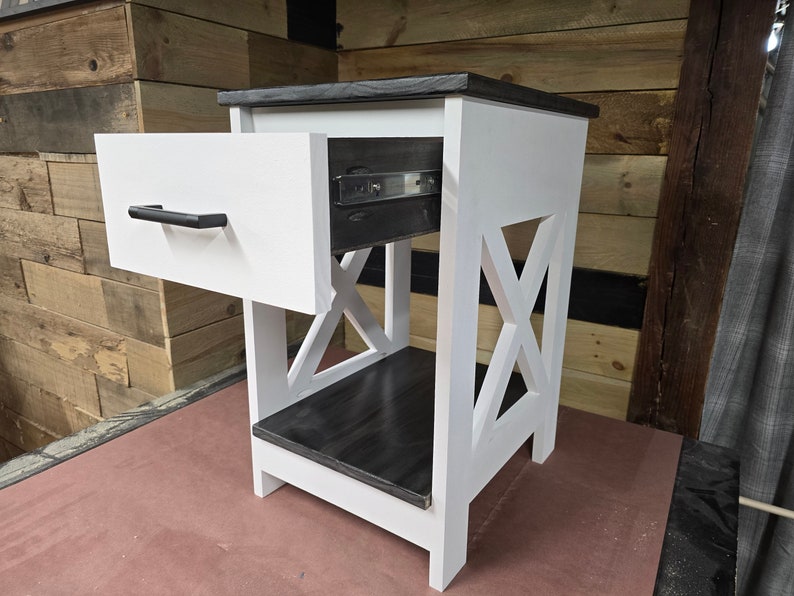 May include: White nightstand with a black top and a single drawer. The drawer is open and has a black pull handle. The nightstand has an X-shaped design on the front.