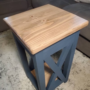 May include: A small wooden end table with a natural wood top and a dark blue painted base. The base has an X-shaped design and a shelf.