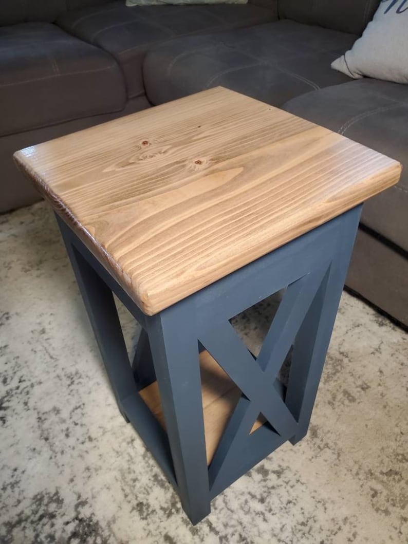 May include: A small wooden end table with a natural wood top and a dark blue painted base. The base has an X-shaped design and a shelf.