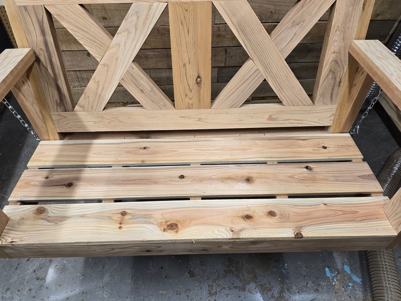 May include: A wooden swing with a slatted seat and a criss-cross design on the back. The swing is made of natural wood and has a rustic look.