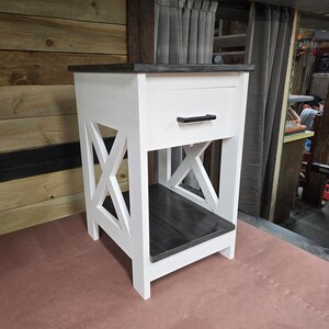 May include: White nightstand with a single drawer and a black drawer pull. The nightstand has a black stained top and bottom shelf. The nightstand has an X-shaped design on the front.