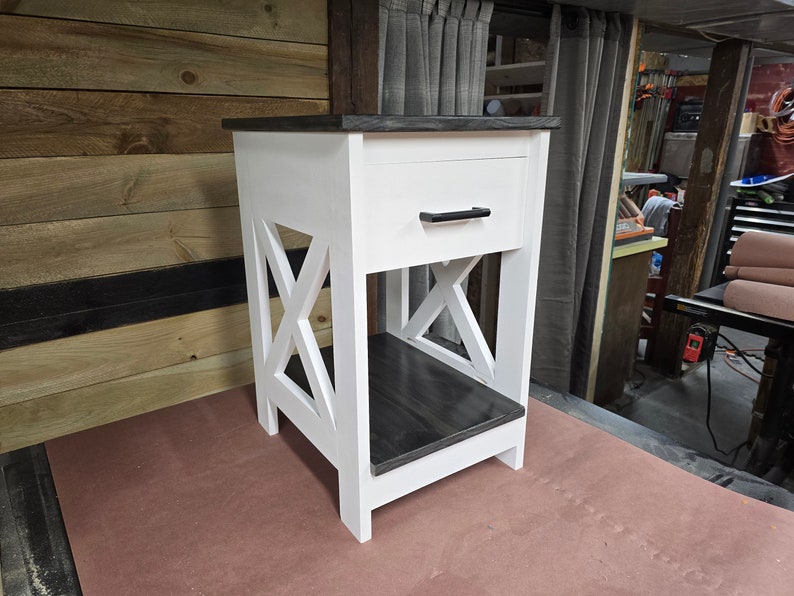 May include: White nightstand with a single drawer and a black drawer pull. The nightstand has a black stained top and bottom shelf. The nightstand has an X-shaped design on the front.