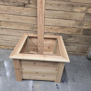 May include: A square wooden planter box with a tall wooden post extending from the center. The planter is made of unfinished wood and has a natural wood color.