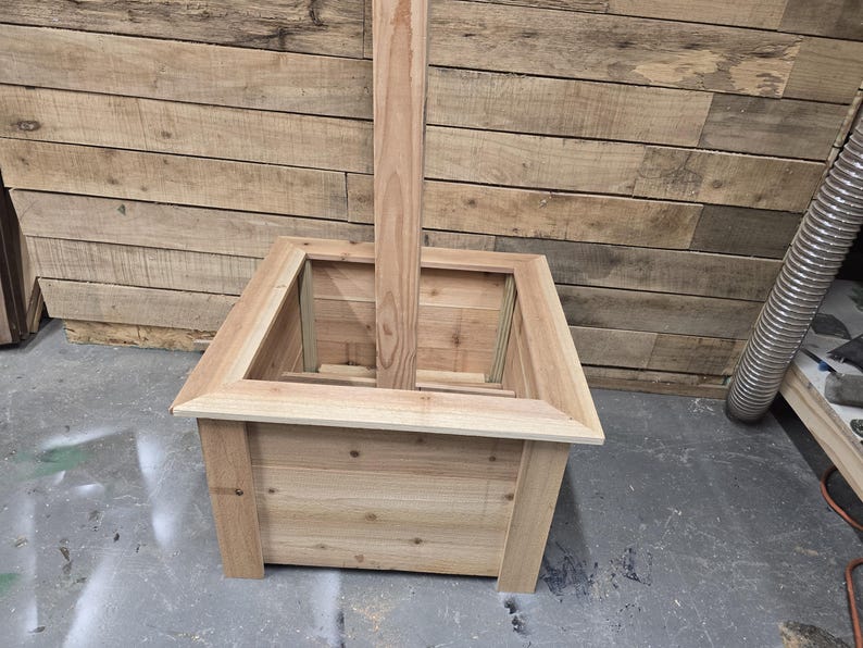 May include: A square wooden planter box with a tall wooden post extending from the center. The planter is made of unfinished wood and has a natural wood color.