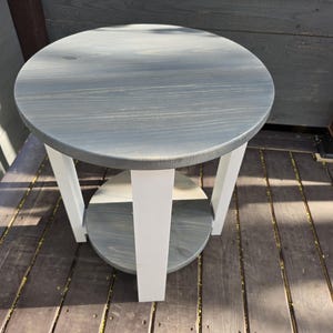 May include: A round wooden end table with a gray stained top and white painted legs. The table has a lower shelf.