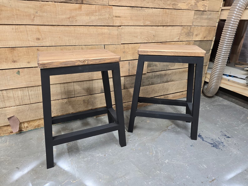 Puede incluir: Dos taburetes de metal negro con tapas de madera. Los taburetes tienen un dise&ntilde;o industrial sencillo y son perfectos para agregar un toque de encanto r&uacute;stico a cualquier espacio.