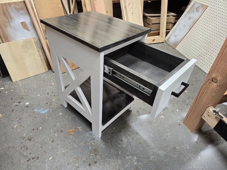 May include: A white and black end table with a drawer that is open. The drawer has a black interior and a black metal slide. The table has a black top and a white base with an X-shaped design.