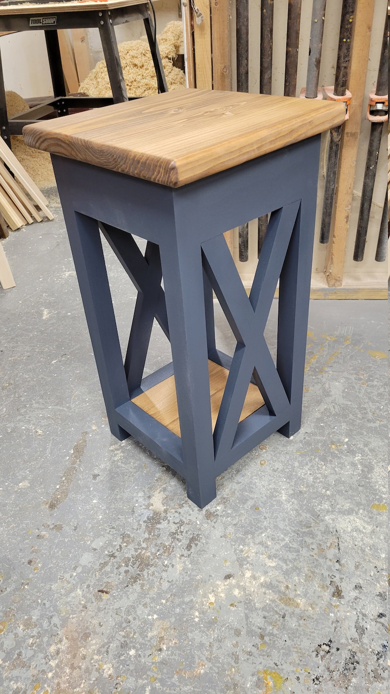 May include: A small, dark blue end table with a wooden top and a shelf. The table has an X-shaped design on the front and sides.