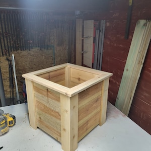 K&ouml;nnte beinhalten: Ein quadratischer Holz-Pflanzkasten aus unbehandeltem hellbraunem Holz. Der Pflanzkasten steht auf einer wei&szlig;en Oberfl&auml;che.