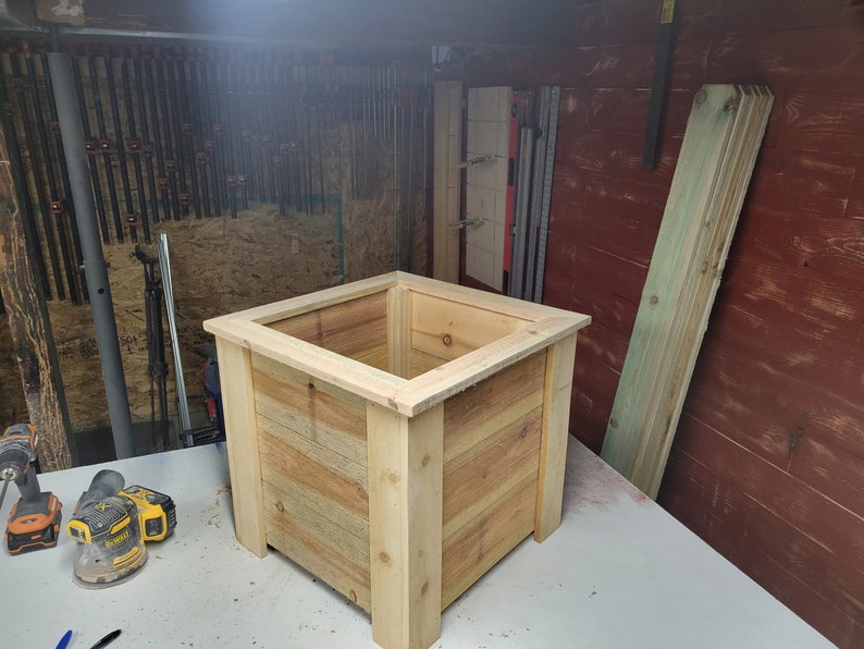 K&ouml;nnte beinhalten: Ein quadratischer Holz-Pflanzkasten aus unbehandeltem hellbraunem Holz. Der Pflanzkasten steht auf einer wei&szlig;en Oberfl&auml;che.