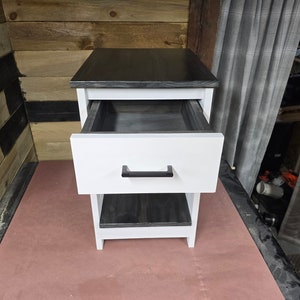 May include: A white nightstand with a black top and a single drawer. The drawer is open and the inside is black. The nightstand has a lower shelf that is also black.