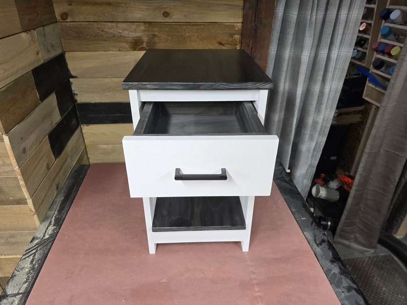 May include: A white nightstand with a black top and a single drawer. The drawer is open and the inside is black. The nightstand has a lower shelf that is also black.
