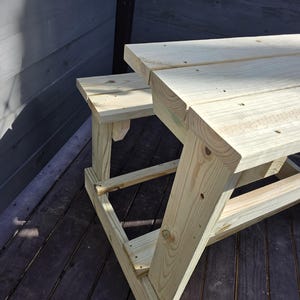 May include: A wooden picnic table with a bench attached to one side. The table and bench are made of unfinished light brown wood.