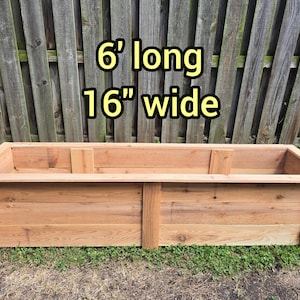 May include: A rectangular wooden planter box with a natural light brown finish, featuring vertical support beams along the sides and a flat top rim. The text 6' long 16" wide is printed above the planter, indicating its dimensions.