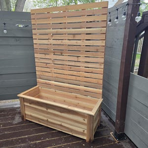 以下が含まれることがあります： 背後に高いスラット付きのプライバシー画面がある木製プランターボックス。プランターボックスは天然のヒノキ材で作られており、長方形です。プライバシー画面もヒノキ材で作られており、スラットデザインになっています。