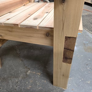 May include: Close-up of a wooden chair with a natural finish. The chair has a simple design with a wide seat and a sturdy frame. The chair is made of solid wood and has a rustic look.