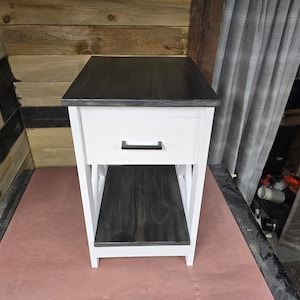 May include: A white end table with a black top and a single drawer. The drawer has a black handle. The table has an open shelf below the drawer.