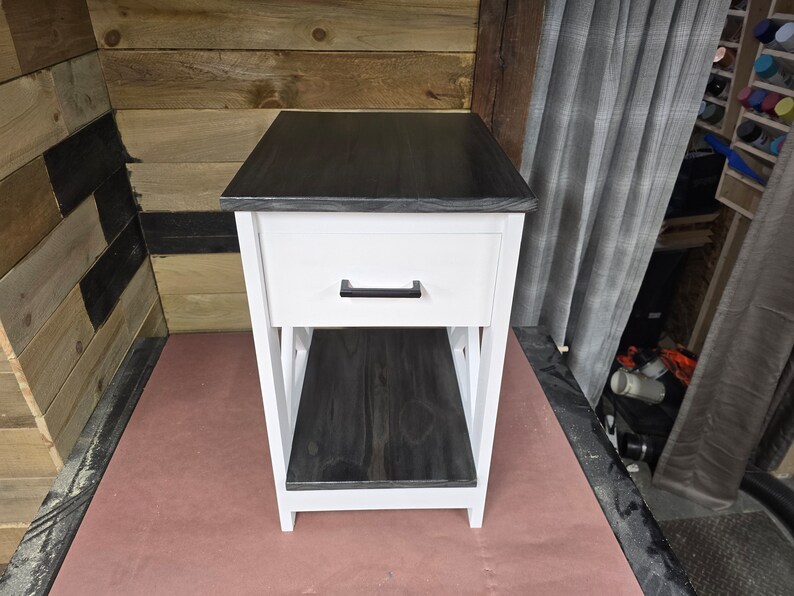 May include: A white end table with a black top and a single drawer. The drawer has a black handle. The table has an open shelf below the drawer.