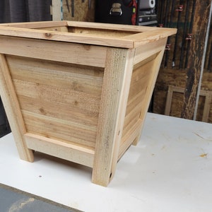 May include: A wooden planter box with a square top and a slightly tapered body. The planter is made of light brown wood and has a natural finish.