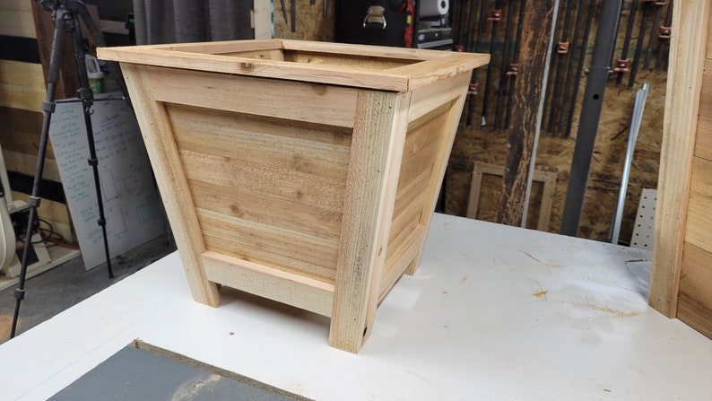 May include: A wooden planter box with a square top and a slightly tapered body. The planter is made of light brown wood and has a natural finish.