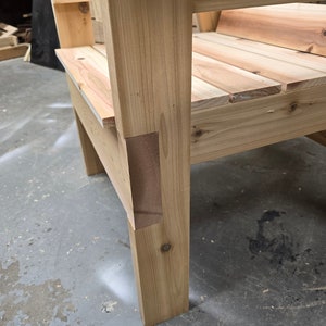 May include: Close-up of a wooden chair with a natural finish. The chair has a simple design with a wide seat and a back that is slightly angled. The chair is made of solid wood and has a sturdy construction.