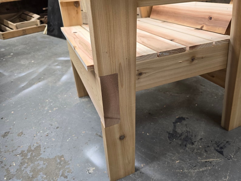 May include: Close-up of a wooden chair with a natural finish. The chair has a simple design with a wide seat and a back that is slightly angled. The chair is made of solid wood and has a sturdy construction.