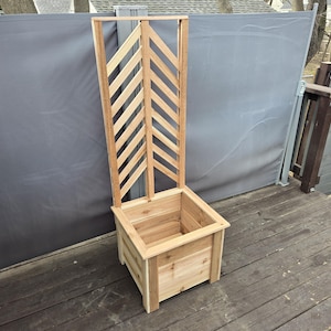 May include: A wooden planter box with a tall, decorative trellis. The planter is rectangular with a natural wood finish. The trellis features a chevron pattern. The structure is designed for outdoor use and is placed on a wooden deck.