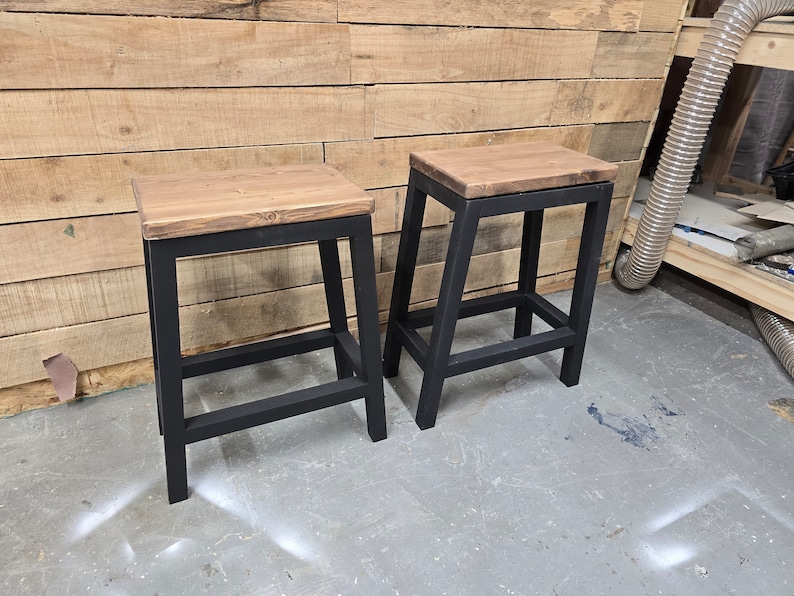 Puede incluir: Dos taburetes de metal negro con tapas de madera. Los taburetes tienen un dise&ntilde;o sencillo con un solo estante debajo del asiento.
