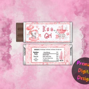 以下が含まれることがあります： ピンクと白のチョコレートバーの包装紙。ピンクの象とベビーカーが描かれています。包装紙には「It's a Girl」と書かれており、栄養成分表も記載されています。