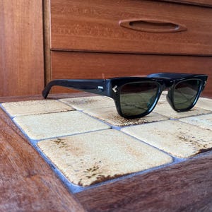 May include: Black sunglasses with a thick frame and green-tinted lenses rest on a tiled surface. The sunglasses have a small, decorative arrow detail on the temples. The background features a wooden cabinet.