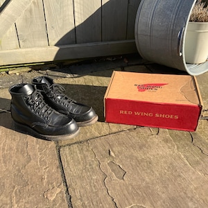 Könnte beinhalten: Ein Paar schwarze Lederschuhe von Red Wing mit Schnürsenkeln. Die Schuhe stehen auf einer Steinoberfläche neben einem roten und braunen Karton mit der Aufschrift "Red Wing Shoes".