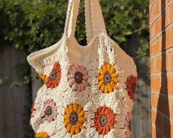 May include: A crocheted tote bag with a cream background and colorful floral designs. The bag has two straps and is made with a granny square pattern.