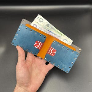 May include: A blue leather wallet with a brown leather flap and a gray leather accent. The wallet has a row of silver rivets around the edges and a small logo on the bottom right corner. The wallet is open and has a few bills and cards inside.