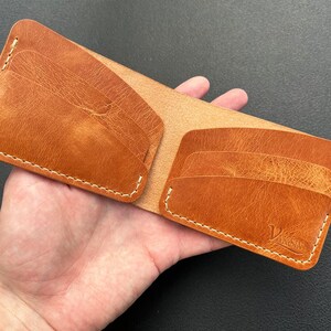 Puede incluir: Una cartera de cuero marrón con múltiples ranuras para tarjetas y un borde cosido. La cartera tiene el texto "V&N" grabado en la parte delantera.