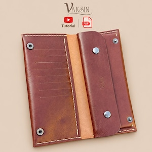 May include: A brown leather long wallet featuring a flap closure with two silver-tone metal snap buttons, white contrast stitching, and multiple interior card slots. The brand name Vaksin is embossed on the interior flap, with tutorial icons above.