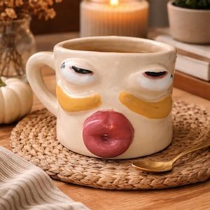 Peut inclure: Une tasse en céramique beige avec un motif de visage. La tasse présente de grands yeux, des accents de joues jaunes et un motif de lèvres rouges. La tasse repose sur un dessous de verre tissé avec une cuillère dorée à proximité.
