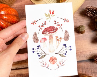 Puede incluir: Una ilustración en acuarela de setas, bayas y piñas en una tarjeta blanca. La tarjeta está sostenida por una mano, con un fondo de madera y flores secas. La obra de arte presenta una seta roja y blanca con lunares.