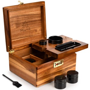 May include: A wooden stash box with an open lid, revealing compartments and a built-in tray. The box features a combination lock, a small brush, and two black jars. The lid is engraved with the word "Moettik" and a logo.