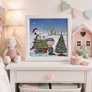 May include: A square white framed print featuring a Peanuts holiday scene with Snoopy wearing a Santa hat on a doghouse, Woodstock, and Charlie Brown standing next to a decorated Christmas tree in a snowy landscape under a starry night sky.