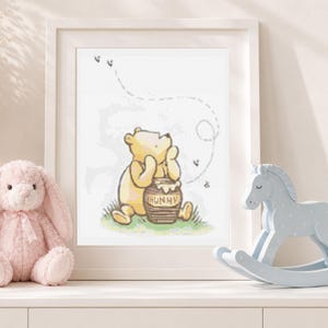 May include: A framed wall art print featuring a yellow bear sitting on green grass next to a brown honey pot labeled HUNNY. The illustration includes a dotted line path of flying bees. The print is displayed in a white frame with a light border.
