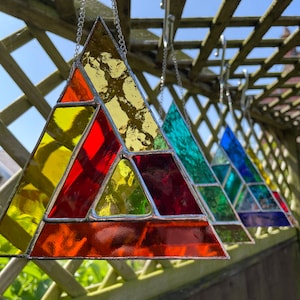 Puede incluir: Un conjunto de cuatro atrapasoles triangulares de vitral colgados de una celosía de madera. Los atrapasoles están hechos de piezas de vidrio de colores en tonos de rojo, naranja, amarillo, verde, azul y morado. Los atrapasoles están suspendidos de cadenas plateadas.