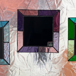 Può includere: Tre specchi quadrati in vetro colorato con cornici nere appese a un muro. Gli specchi hanno pezzi di vetro di diversi colori nelle cornici. Il primo specchio ha pezzi di vetro blu, verde e viola. Il secondo specchio ha pezzi di vetro rosa, viola e bianco. Il terzo specchio ha pezzi di vetro verde, giallo e blu.