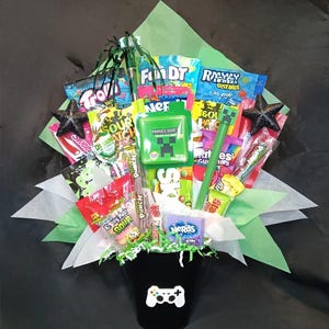 May include: A black bucket with a white game controller icon containing a bouquet of assorted candy including Minecraft, Nerds, Sour Patch Kids, and Skittles. The arrangement is wrapped in light green paper with two black glittery star accents.