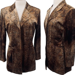 Op de afbeelding: Een bruine blazer met een slangenprint. De blazer heeft een revers, twee voorzakken en een sluiting met één knoop. De stof heeft een textuur en een donkerbruine fluwelen afwerking.