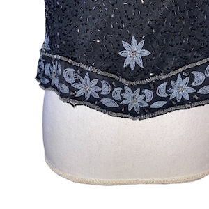 May include: A black sequined top with a silver beaded trim. The trim features a repeating pattern of flowers and crescent moons.