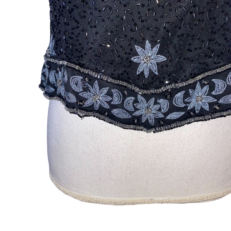 May include: A black sequined top with a silver beaded trim. The trim features a repeating pattern of flowers and crescent moons.